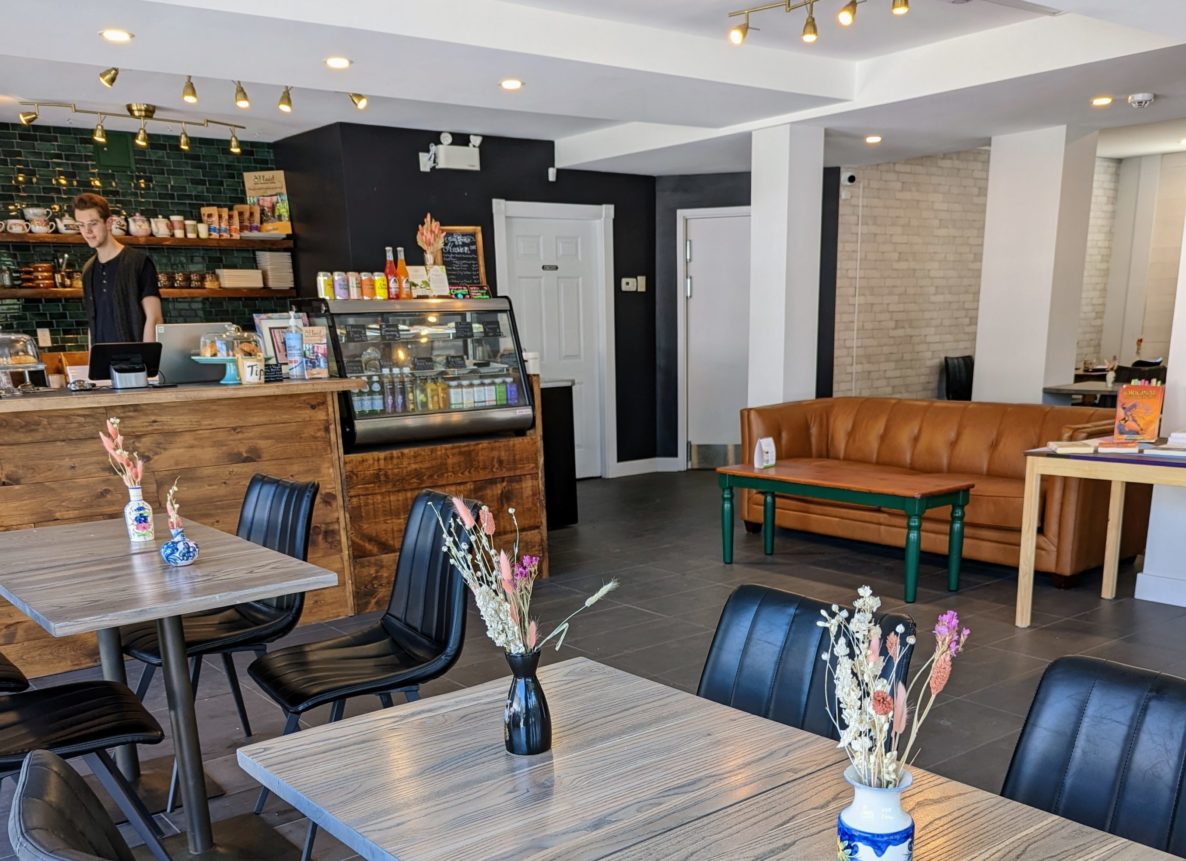 Photo of Haven cafe as seen from the Sunnyside St window. Pictured is several wood tables with vintage black leather chairs, a large tan leather couch and the coffee bar with emerald green tiling and thrift shop mugs.